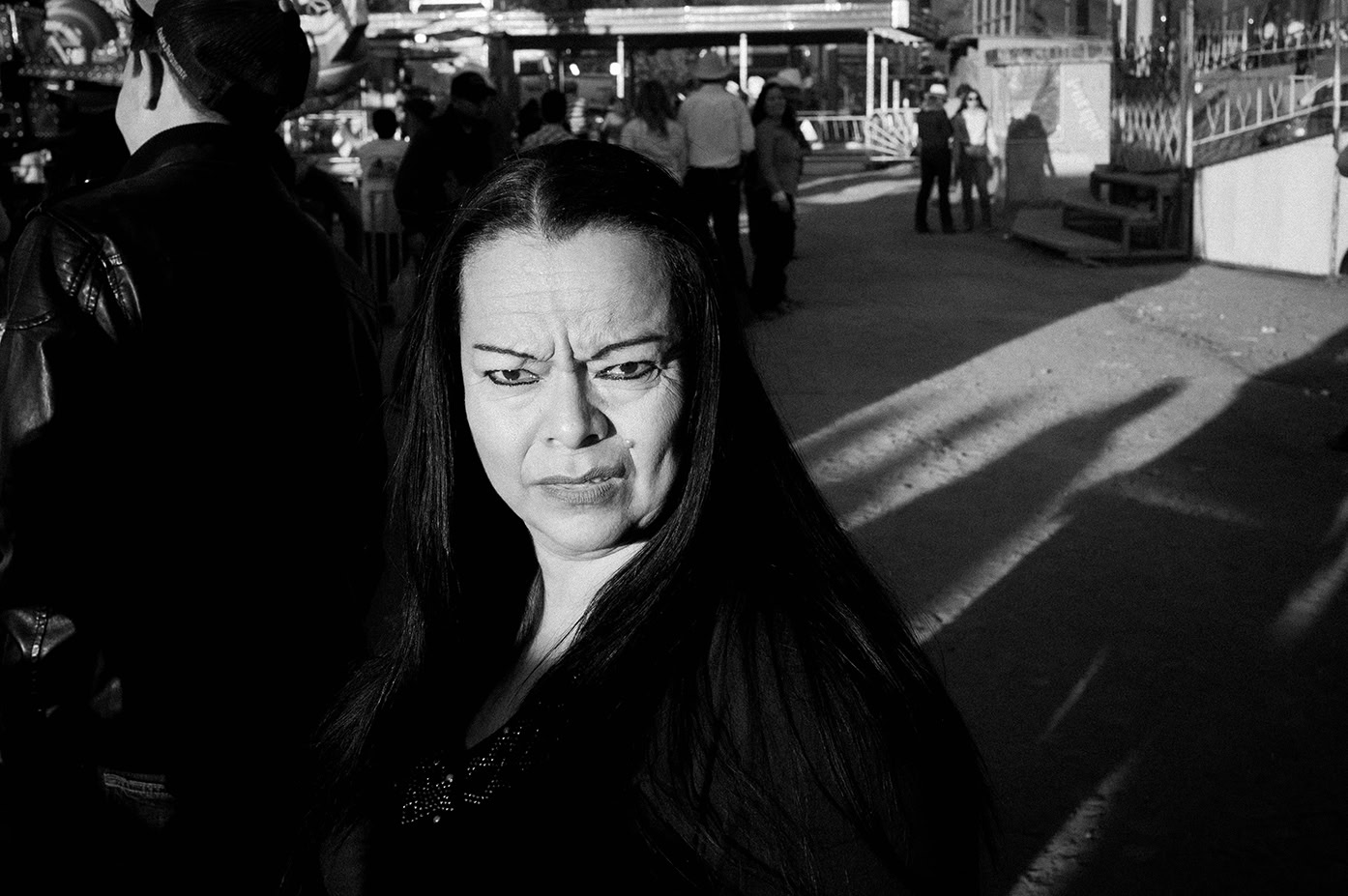 A woman with long dark hair stands outdoors in bright sunlight, looking slightly concerned. Several people and cast shadows are visible in the background.