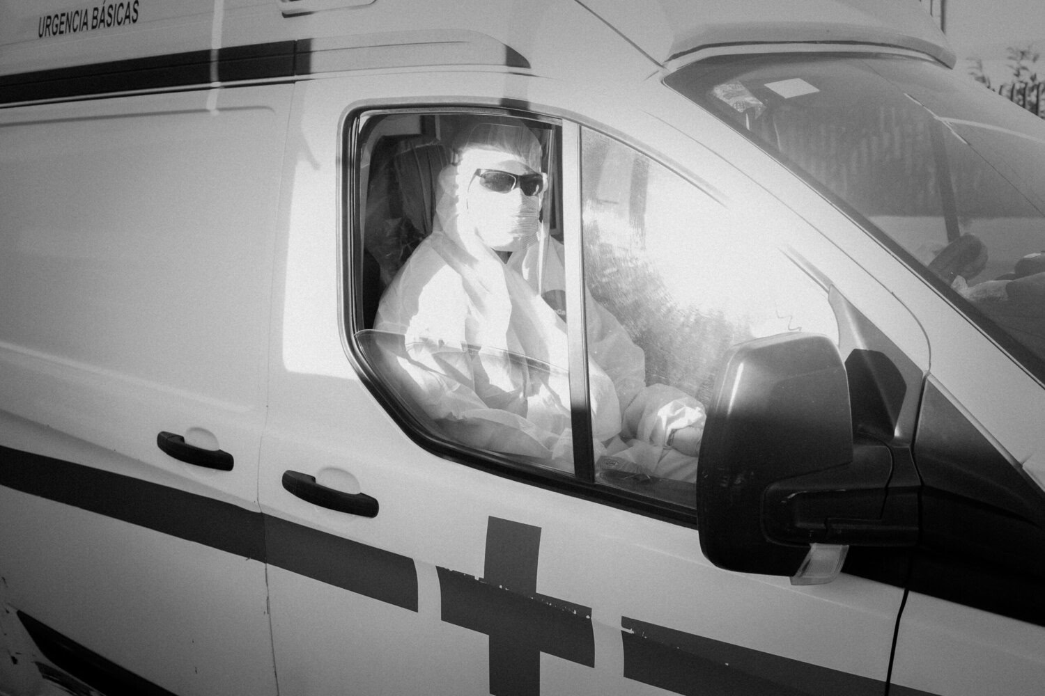 Person wearing protective gear and sunglasses sits in the driver’s seat of an ambulance, viewed through the window, with sunlight reflecting on the glass.