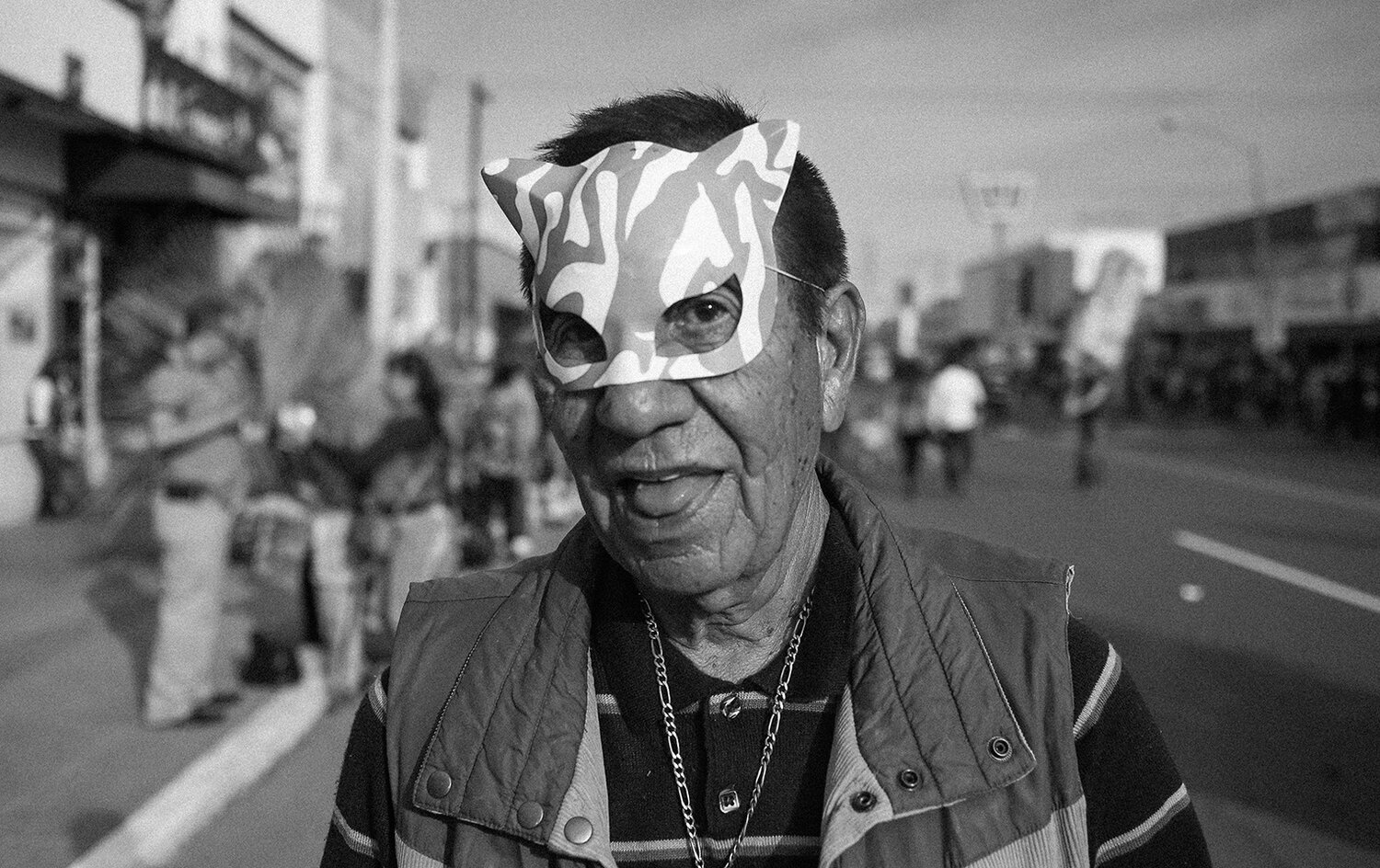 An older man wearing a zebra-patterned mask and a vest stands on a street during what appears to be a public event or parade.
