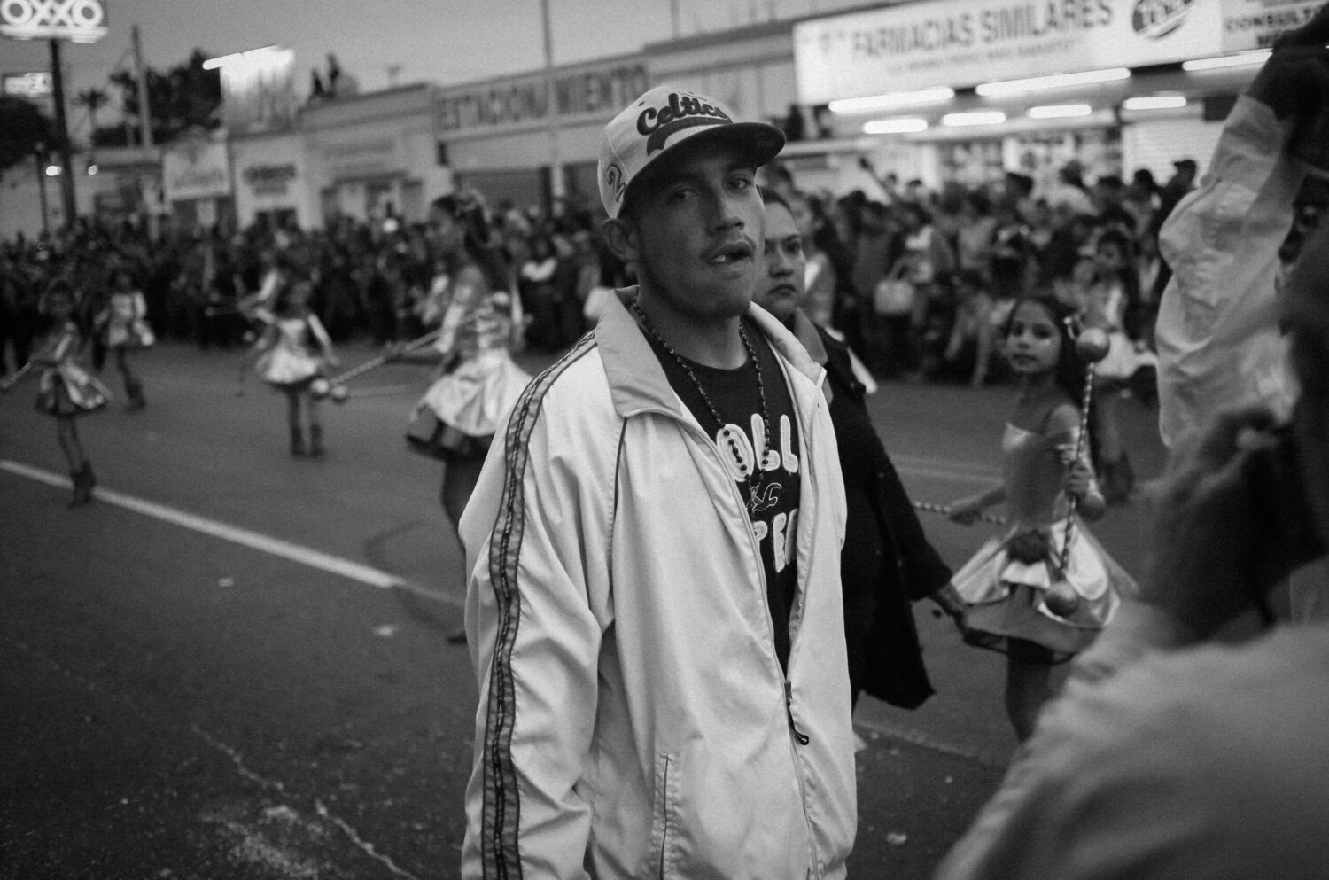 A man in a white jacket and cap walks along a street during a parade, with dancers and onlookers visible in the background.