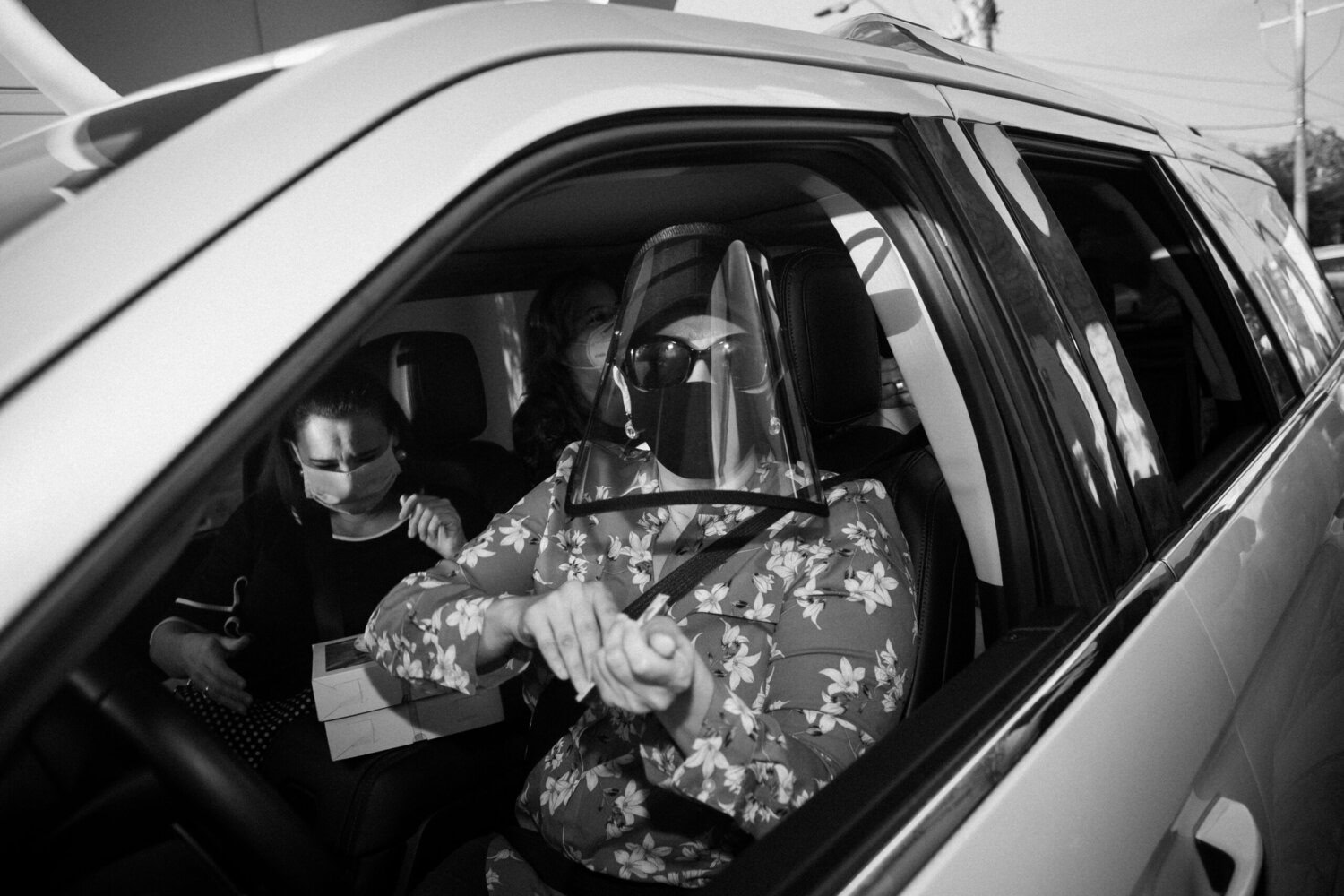 A person in a floral shirt and face shield sits in the driver’s seat of a car, with another masked person in the backseat holding a box.