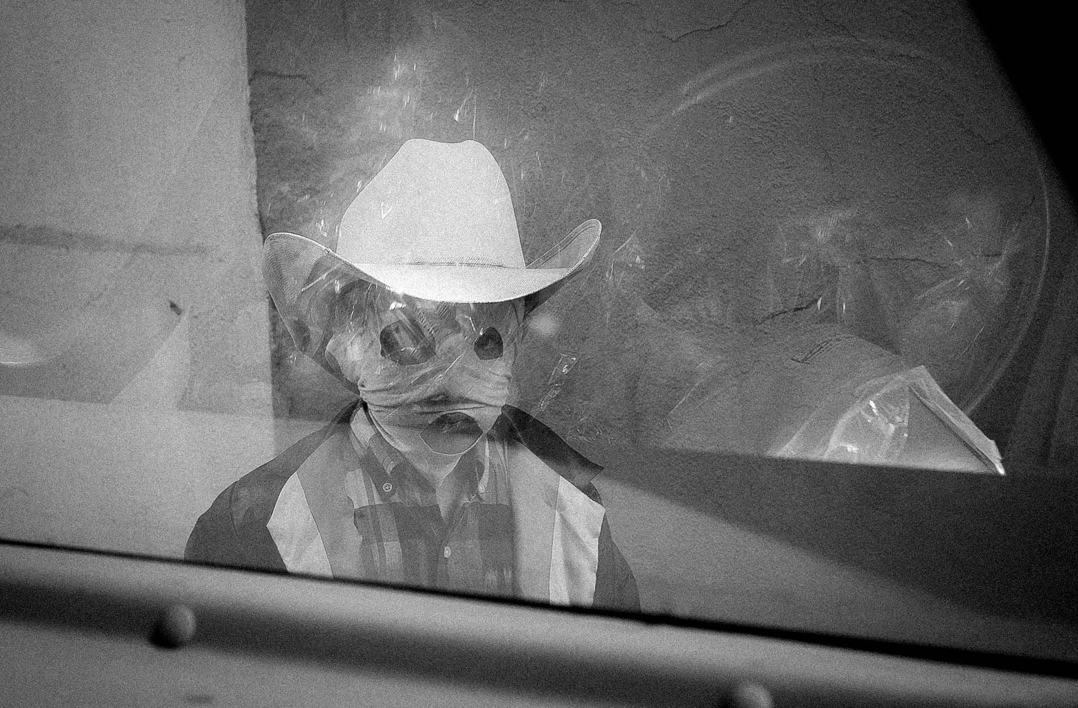A person in a cowboy hat and buttoned shirt stands behind glass, their face obscured by plastic wrap, in a black and white photo.