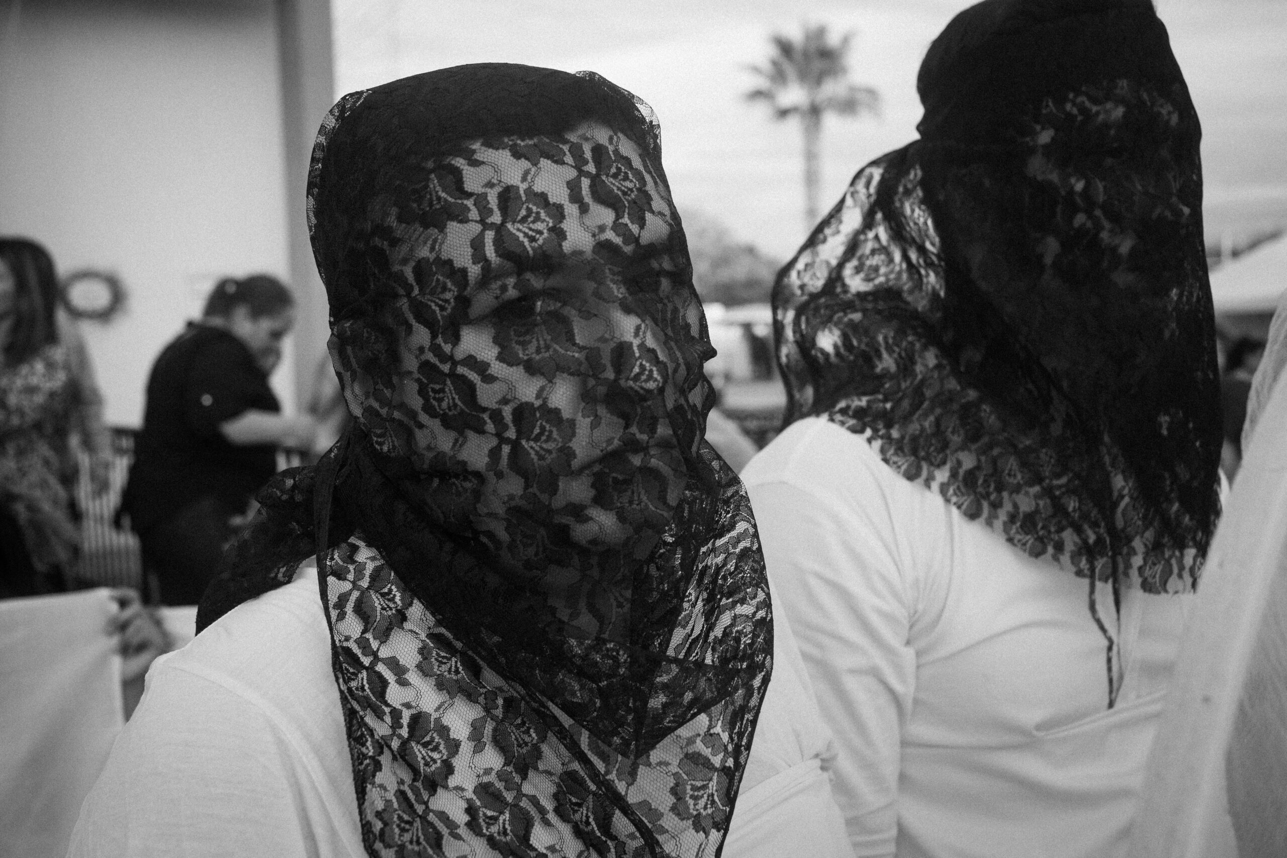 A person wearing a black lace veil over their face stands among others similarly dressed, outdoors in daylight.