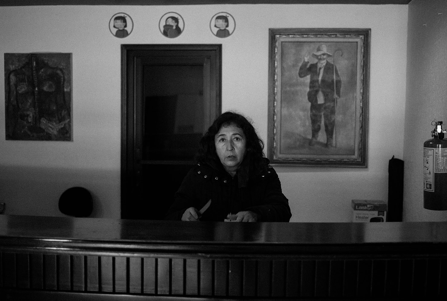 A woman sits behind a wooden desk in a dimly lit room, facing the camera. Framed portraits and a fire extinguisher are visible on the wall behind her.