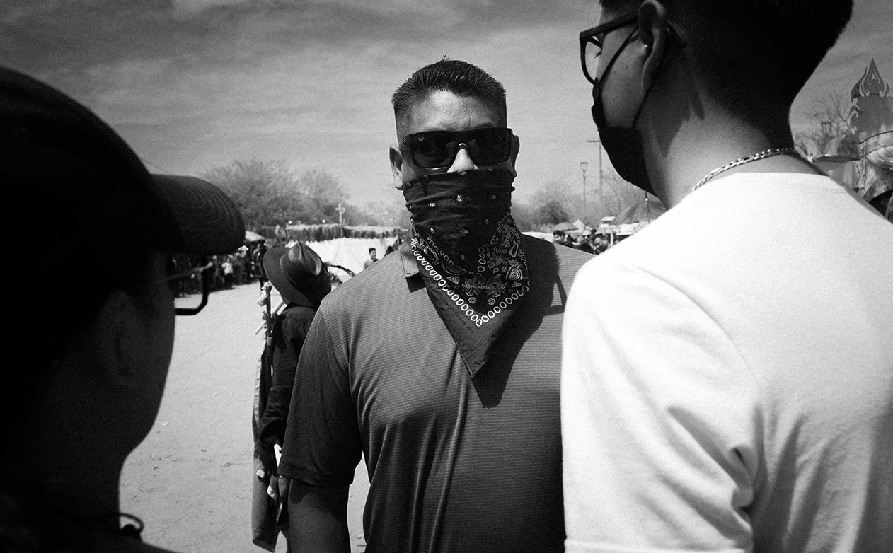 Three people stand outdoors; the central figure wears sunglasses, a bandana over his face, and a t-shirt, while the others wear masks. The background shows a crowd and trees under a bright sky.