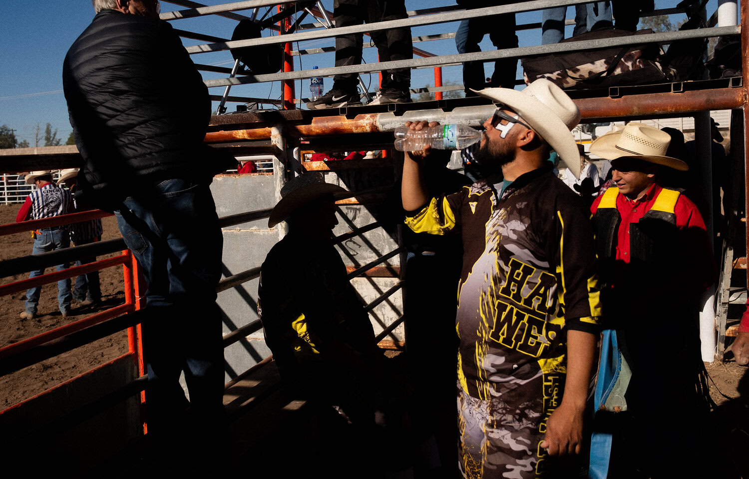 Un hombre con sombrero de vaquero bebe agua junto a una pista de rodeo. Otras personas, vestidas al estilo del Oeste, están de pie o sentadas cerca, algunas a la sombra, detrás de unas barandillas metálicas.