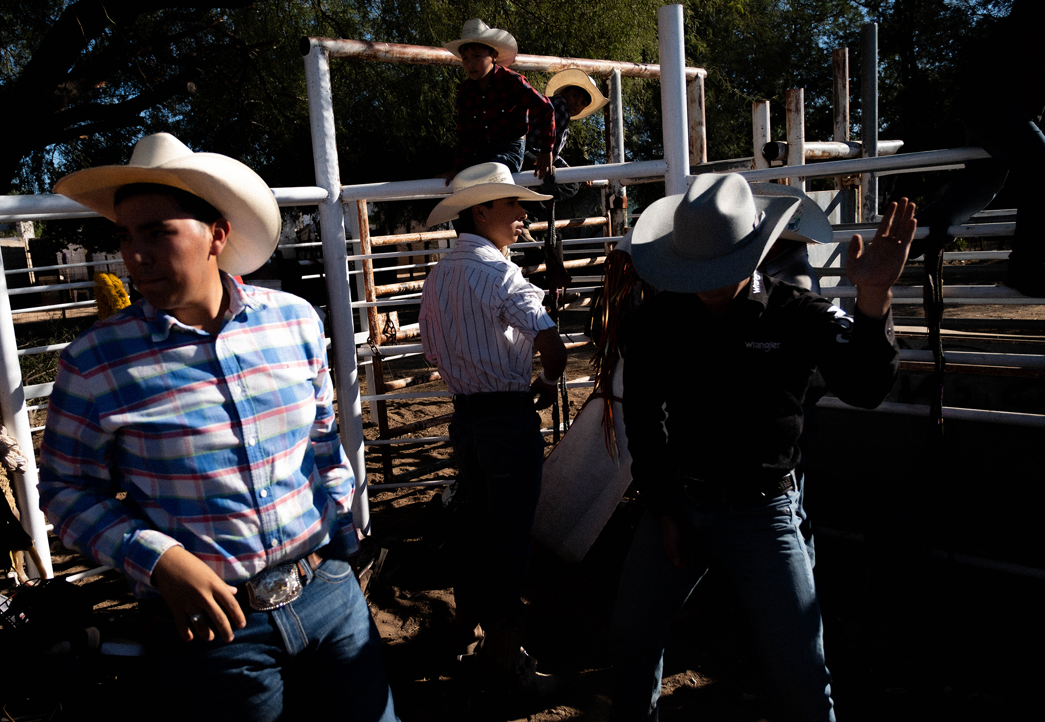 Cuatro personas con sombreros de vaquero están de pie charlando junto a unas barandillas metálicas en un espacio al aire libre, mientras la luz del sol proyecta sombras marcadas sobre la escena.