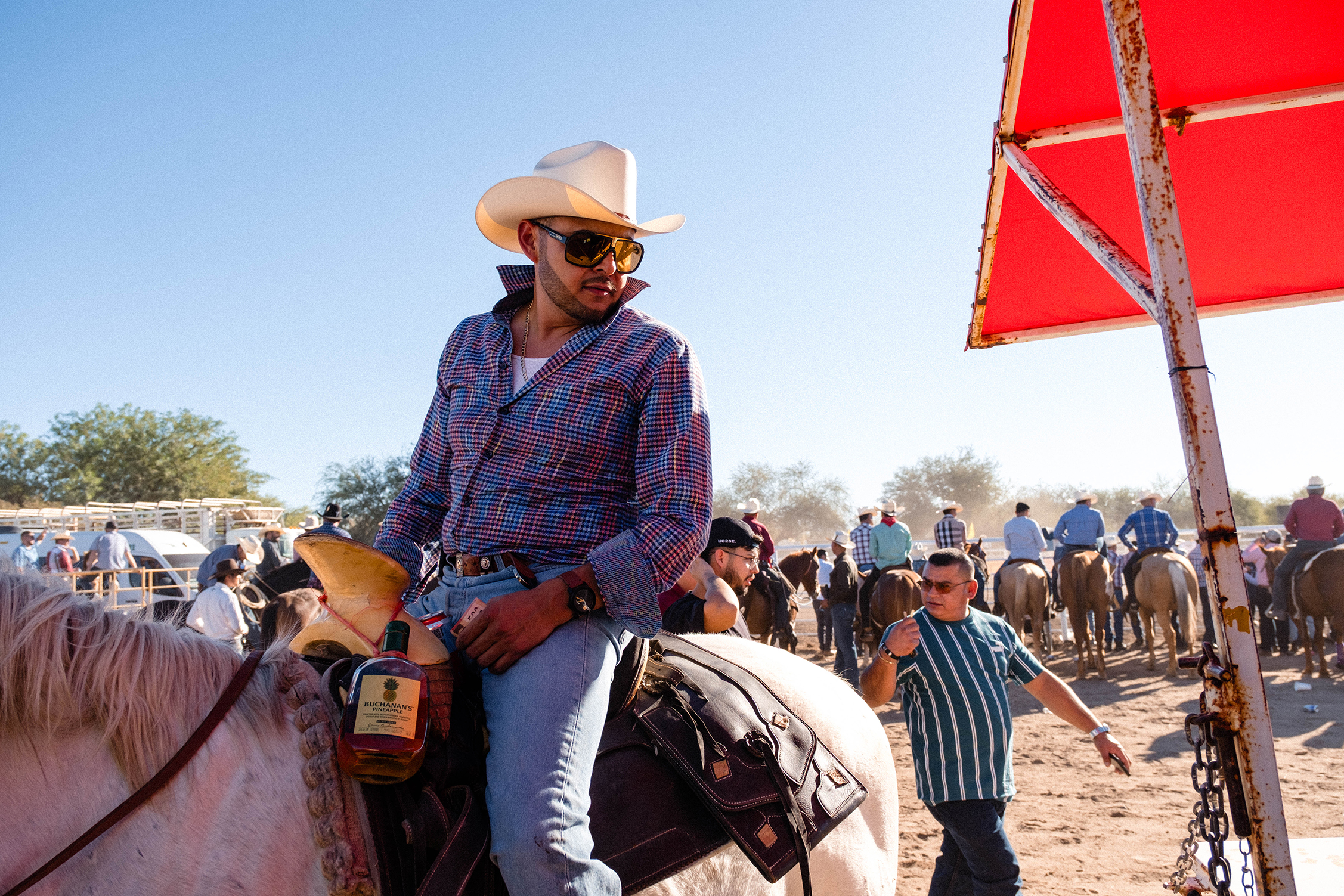 Un hombre con sombrero de vaquero y gafas de sol está sentado sobre un caballo blanco, sosteniendo una botella, con una multitud y caballos al fondo, bajo un cielo azul y despejado.