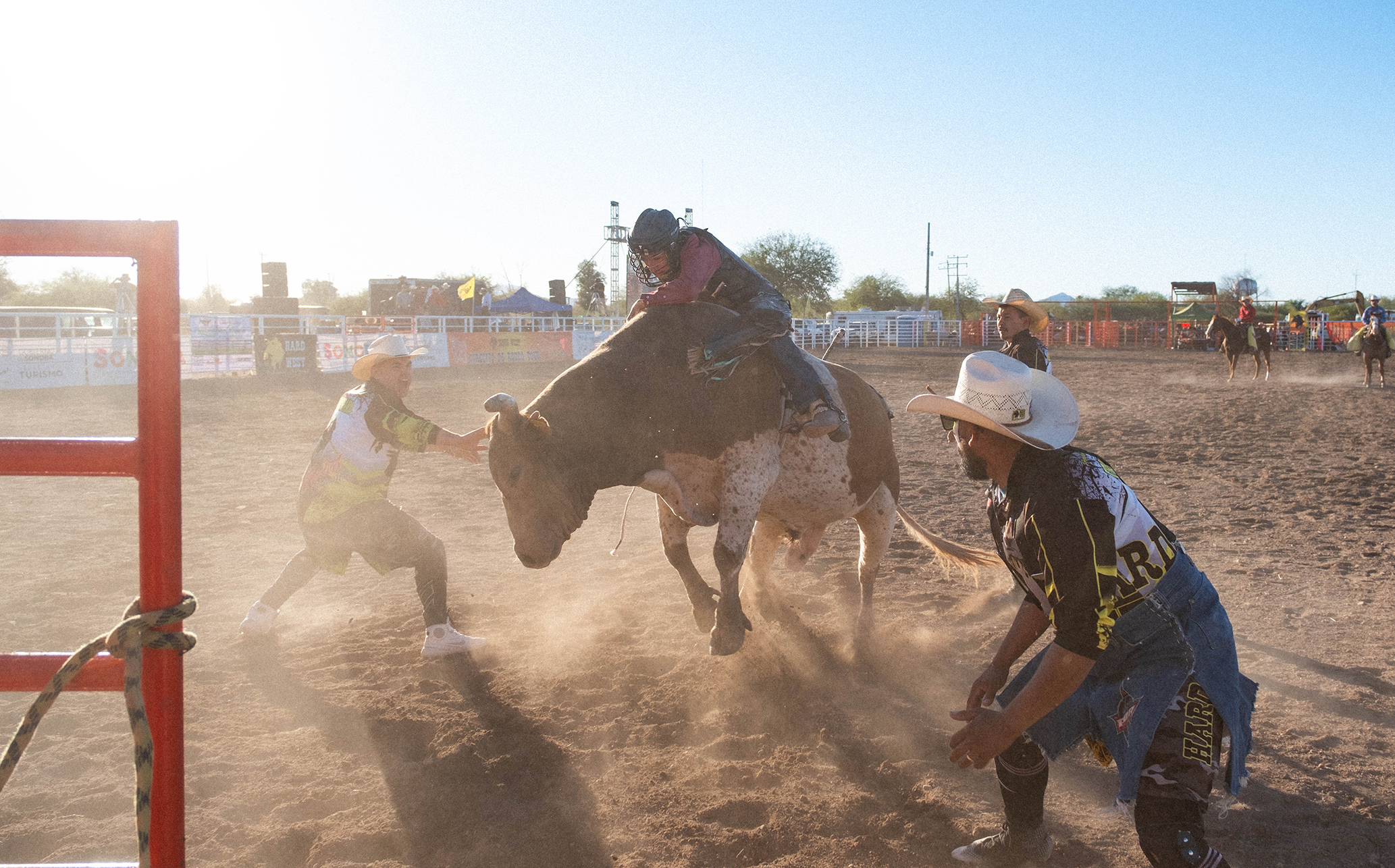 Un jinete de toros se aferra a la bestia mientras dos payasos de rodeo permanecen cerca, listos para ayudarle, en una polvorienta pista al aire libre durante un evento de rodeo.